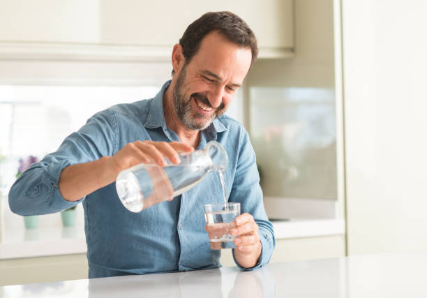 middle age man drinking a glass of water with a happy face standing and smiling with a